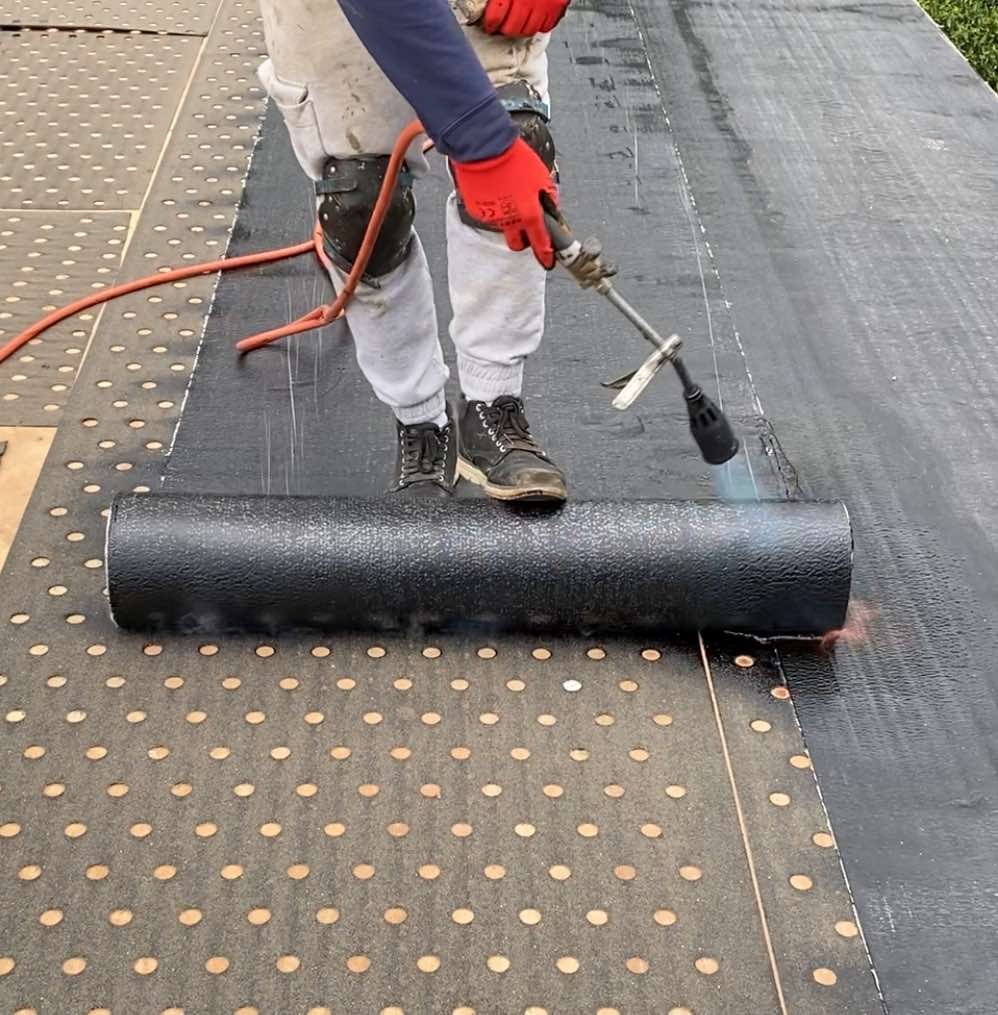 Man spraying black rubber mat onto built-up felt roofing with spray gun.