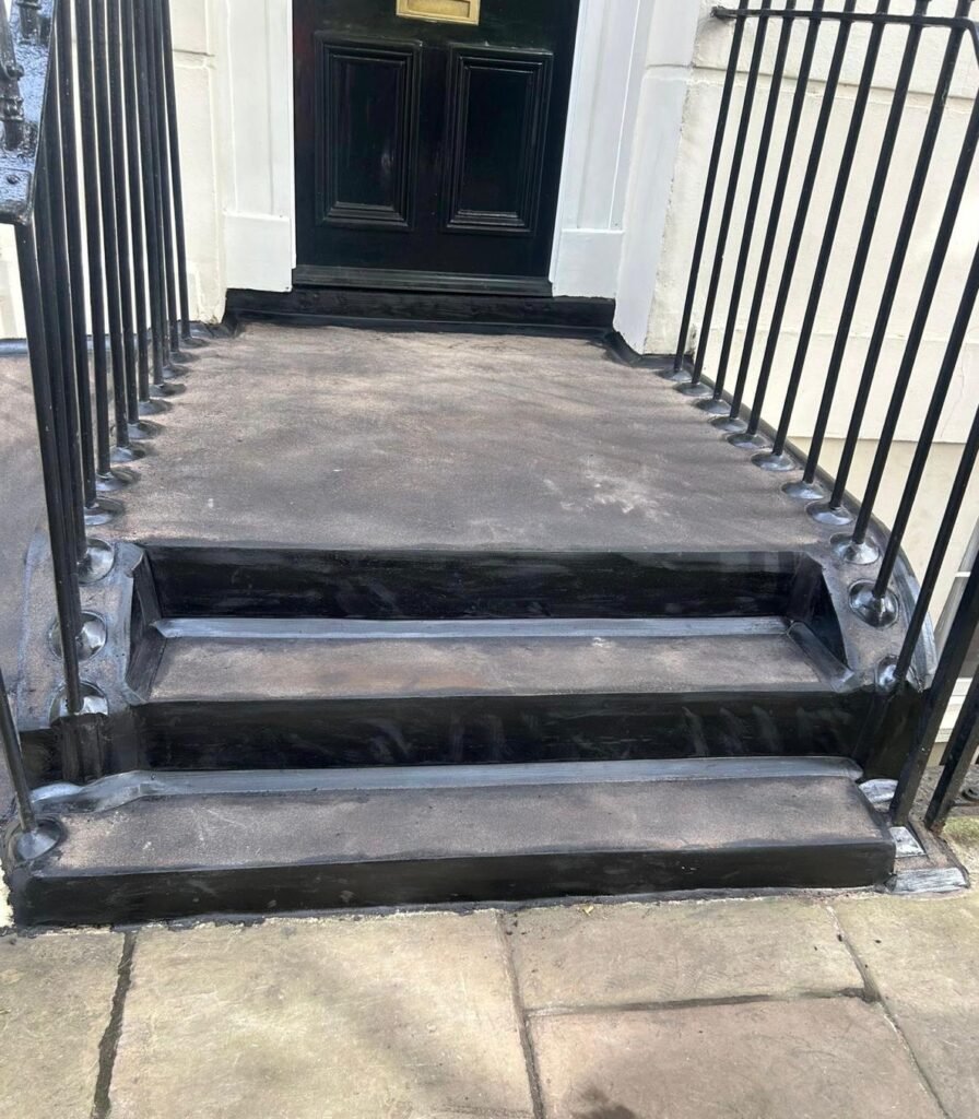 A front porch with black asphalt steps and smooth railings leading up to a black door. The steps are neat and clean with a solid finish.