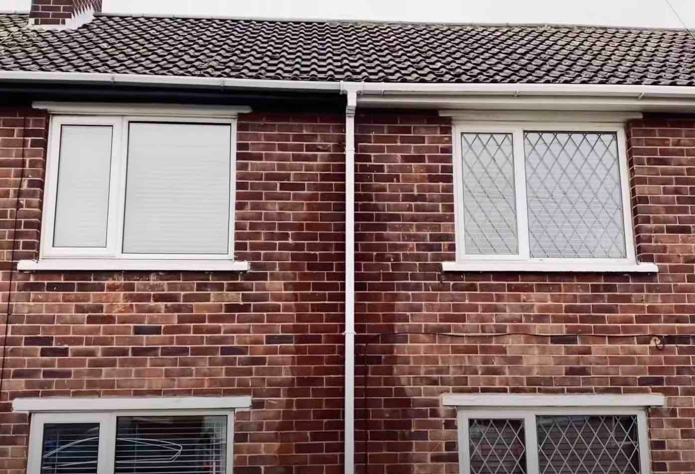 Two windows on a brick house, featuring white frames, with visible signs of a gutter problem above them.