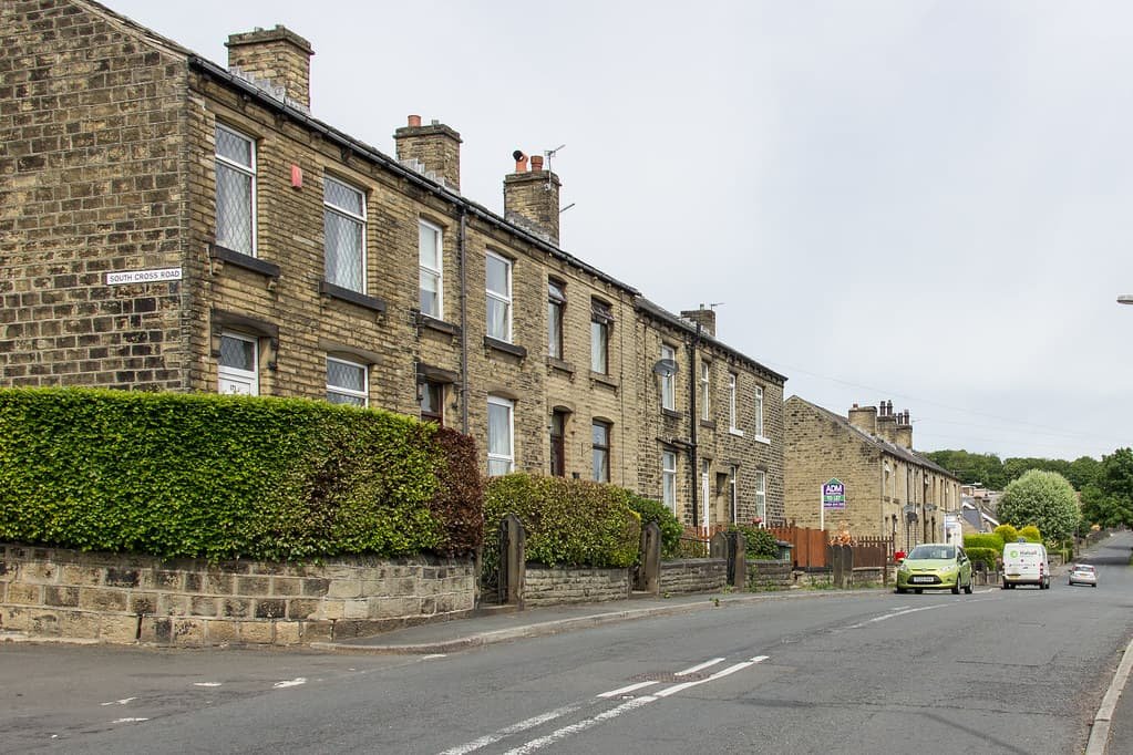 Row of traditional stone houses on a quiet street with green hedges and parked cars.